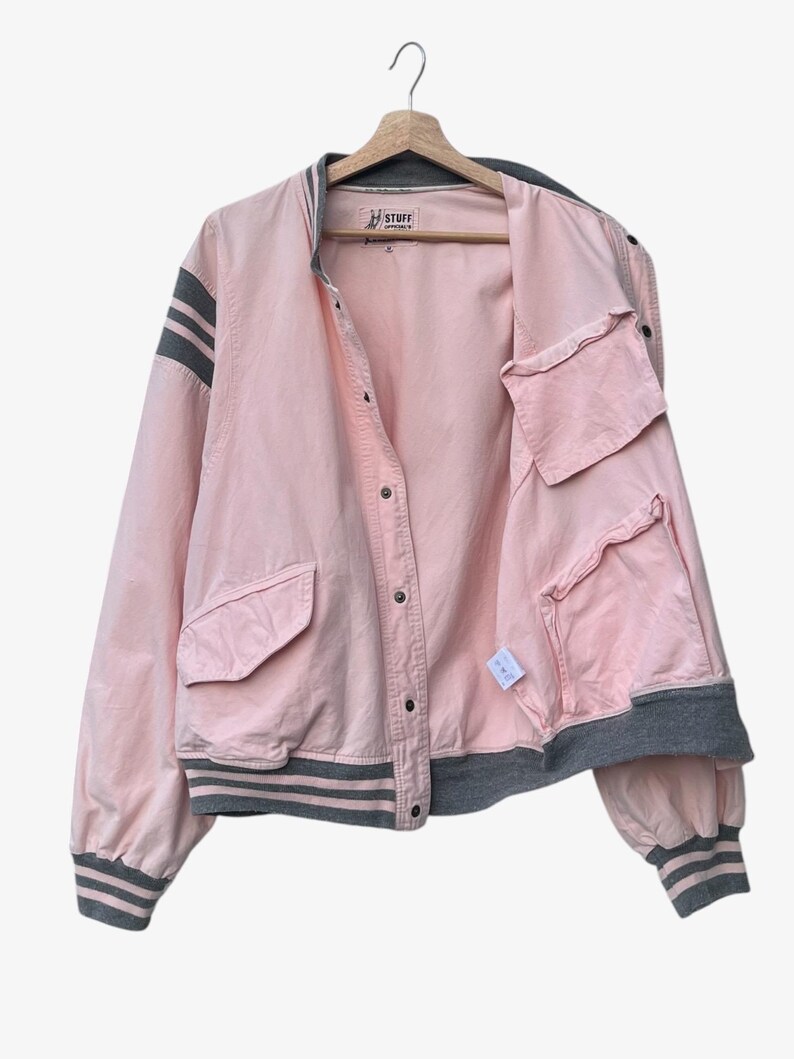 Puede incluir: Chaqueta bomber rosa con detalles grises, incluyendo rayas en las mangas y el ribete. La chaqueta tiene cierre de botones, bolsillos y la palabra "STUFF" en la etiqueta.