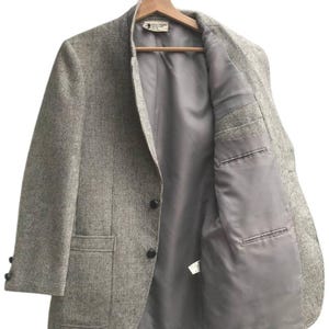 May include: A gray tweed blazer with a two-button closure, two front pockets, and a notched lapel. The blazer is hanging on a wooden hanger, revealing a gray interior lining and a small tag.