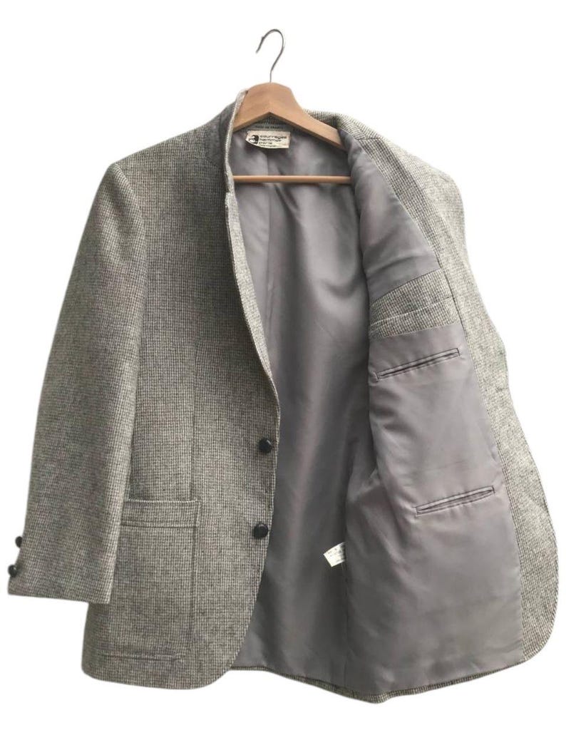 May include: A gray tweed blazer with a two-button closure, two front pockets, and a notched lapel. The blazer is hanging on a wooden hanger, revealing a gray interior lining and a small tag.