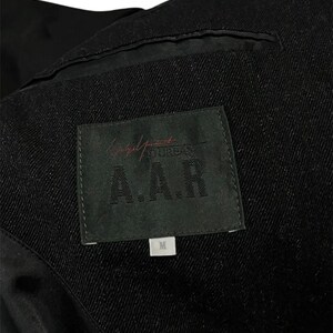 K&ouml;nnte beinhalten: Nahaufnahme des Innenraums einer schwarzen Jacke. Ein schwarzes Etikett mit der Aufschrift "Yohji Yamamoto A.A.R" ist zu sehen, zusammen mit einem kleineren wei&szlig;en Etikett mit der Aufschrift "M". Der Stoff der Jacke hat eine Textur.