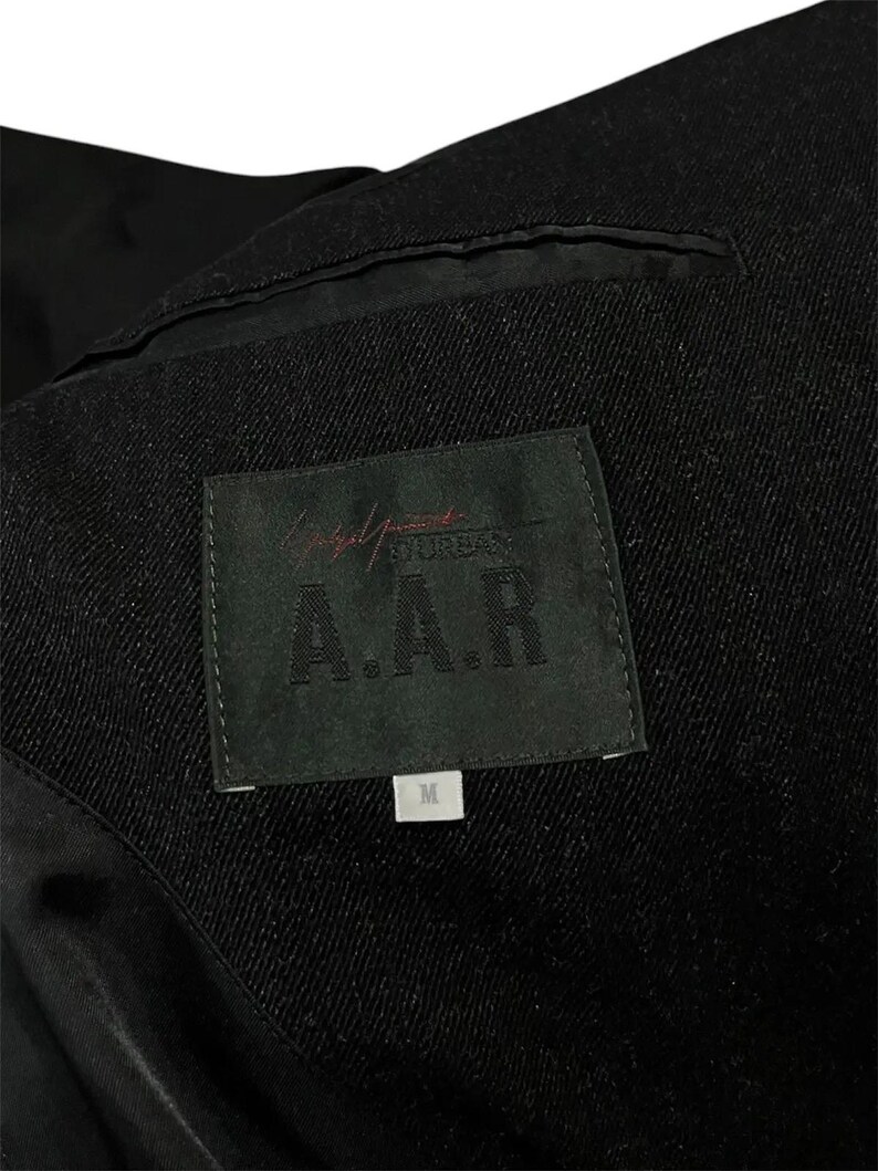 K&ouml;nnte beinhalten: Nahaufnahme des Innenraums einer schwarzen Jacke. Ein schwarzes Etikett mit der Aufschrift "Yohji Yamamoto A.A.R" ist zu sehen, zusammen mit einem kleineren wei&szlig;en Etikett mit der Aufschrift "M". Der Stoff der Jacke hat eine Textur.