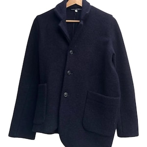 May include: A dark navy wool blazer with a notched lapel, three buttons, and two front pockets. The blazer is hanging on a wooden hanger against a white background. The jacket has a unique asymmetrical hemline.