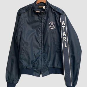 May include: A navy blue Atari jacket with a white Atari logo on the chest and the word "ATARI" printed vertically on the right sleeve. The jacket has a zip-up front, a stand-up collar, and elastic cuffs and waistband.