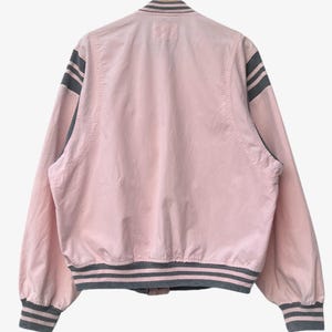 Puede incluir: Chaqueta bomber rosa claro con detalles grises. La chaqueta presenta rayas grises en las mangas, el cuello y el dobladillo. Cuelga de una percha de madera sobre un fondo blanco.