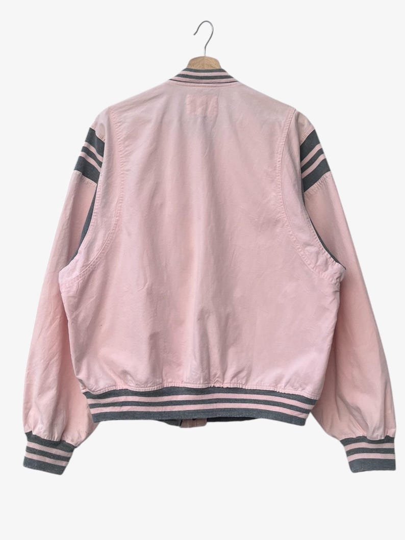 Puede incluir: Chaqueta bomber rosa claro con detalles grises. La chaqueta presenta rayas grises en las mangas, el cuello y el dobladillo. Cuelga de una percha de madera sobre un fondo blanco.