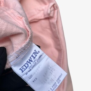 Puede incluir: Primer plano de una prenda rosa claro con una etiqueta blanca. La etiqueta muestra la marca "EDWIN" e incluye informaci&oacute;n sobre el cuidado y la composici&oacute;n de la fibra. Se aprecia la textura y el color de la tela.