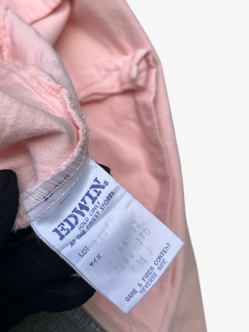 Puede incluir: Primer plano de una prenda rosa claro con una etiqueta blanca. La etiqueta muestra la marca "EDWIN" e incluye informaci&oacute;n sobre el cuidado y la composici&oacute;n de la fibra. Se aprecia la textura y el color de la tela.