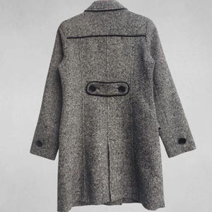 May include: A gray tweed coat with black trim and buttons. The coat has a classic design with a notched collar, long sleeves, and a button closure. The back of the coat features decorative black trim and two buttons. The coat is hanging on a wooden hanger.