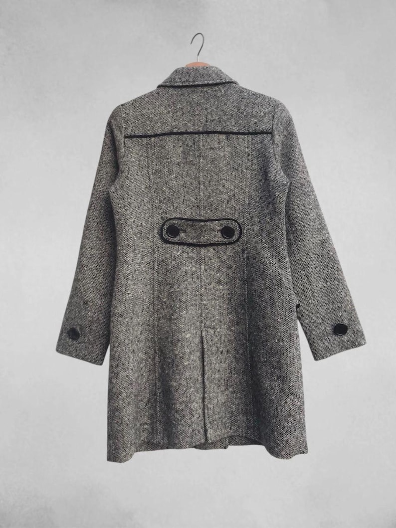 May include: A gray tweed coat with black trim and buttons. The coat has a classic design with a notched collar, long sleeves, and a button closure. The back of the coat features decorative black trim and two buttons. The coat is hanging on a wooden hanger.