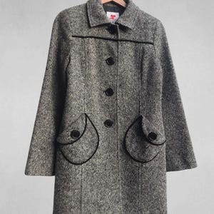 May include: A gray tweed coat with black trim and buttons, hanging on a wooden hanger. The coat features a classic collar, three buttons down the front, and two rounded pockets with black trim.