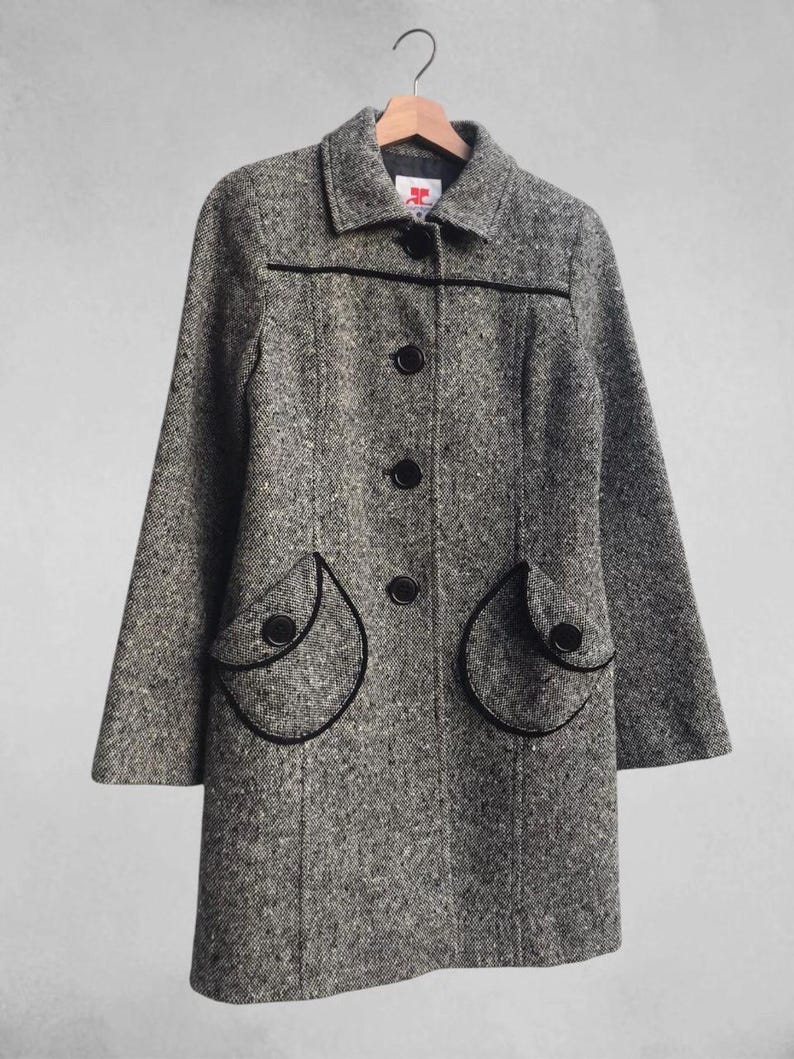 May include: A gray tweed coat with black trim and buttons, hanging on a wooden hanger. The coat features a classic collar, three buttons down the front, and two rounded pockets with black trim.
