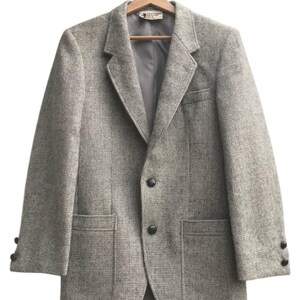 May include: A gray tweed blazer with a two-button closure, notched lapels, and two front pockets. The jacket is hanging on a wooden hanger. The sleeves have three buttons at the cuffs.