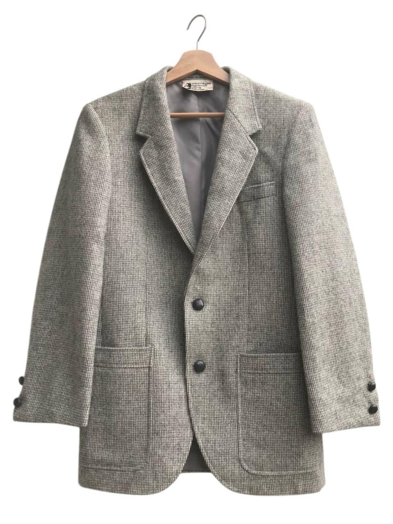 May include: A gray tweed blazer with a two-button closure, notched lapels, and two front pockets. The jacket is hanging on a wooden hanger. The sleeves have three buttons at the cuffs.