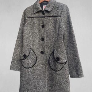 May include: A gray tweed coat with black trim and buttons. The coat has two decorative pockets with black trim and a classic collar. The coat is hanging on a wooden hanger.