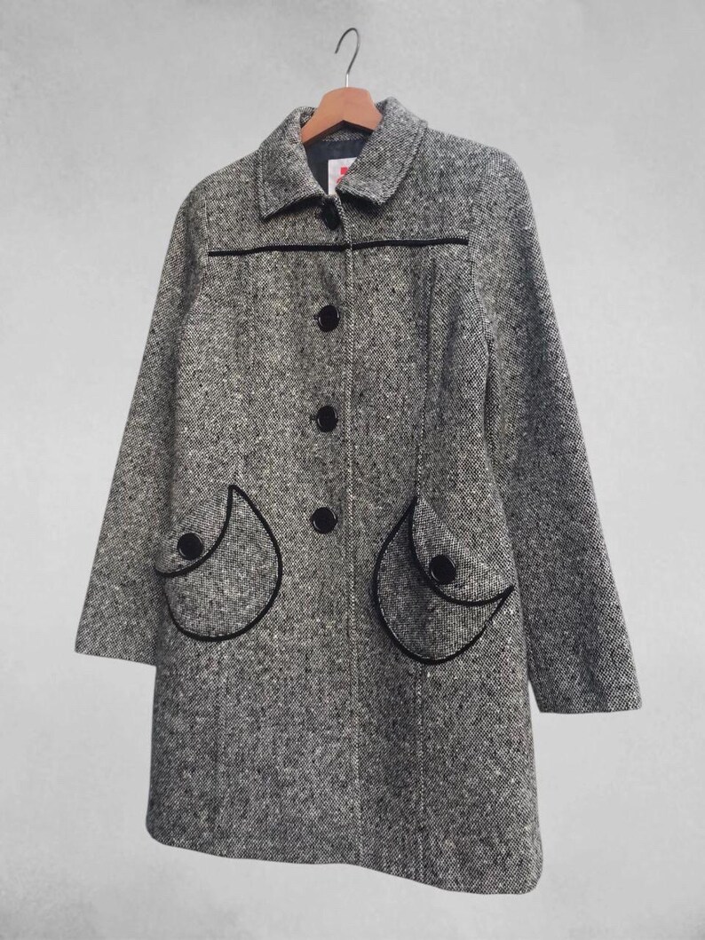 May include: A gray tweed coat with black trim and buttons. The coat has two decorative pockets with black trim and a classic collar. The coat is hanging on a wooden hanger.