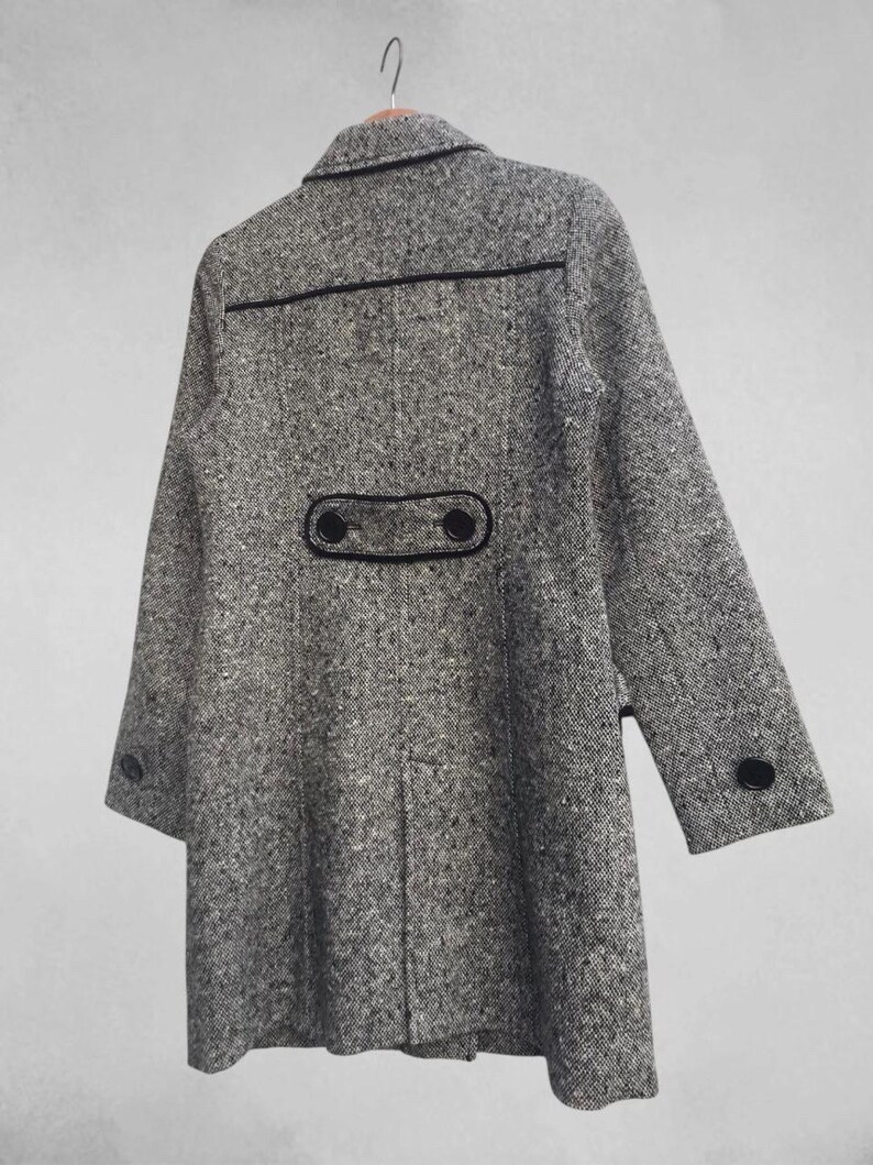 May include: A gray tweed coat with black trim and buttons. The coat is hanging on a wooden hanger. The coat has a classic design with a notched collar and two rows of buttons. The coat is made of a textured fabric.