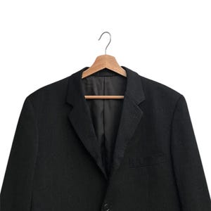 K&ouml;nnte beinhalten: Ein schwarzer Blazer h&auml;ngt an einem Holzkleiderb&uuml;gel. Der Blazer hat ein fallendes Revers, einen Ein-Knopf-Verschluss und eine Brusttasche. Der Stoff ist einfarbig dunkel und das Kleidungsst&uuml;ck ist ma&szlig;geschneidert.