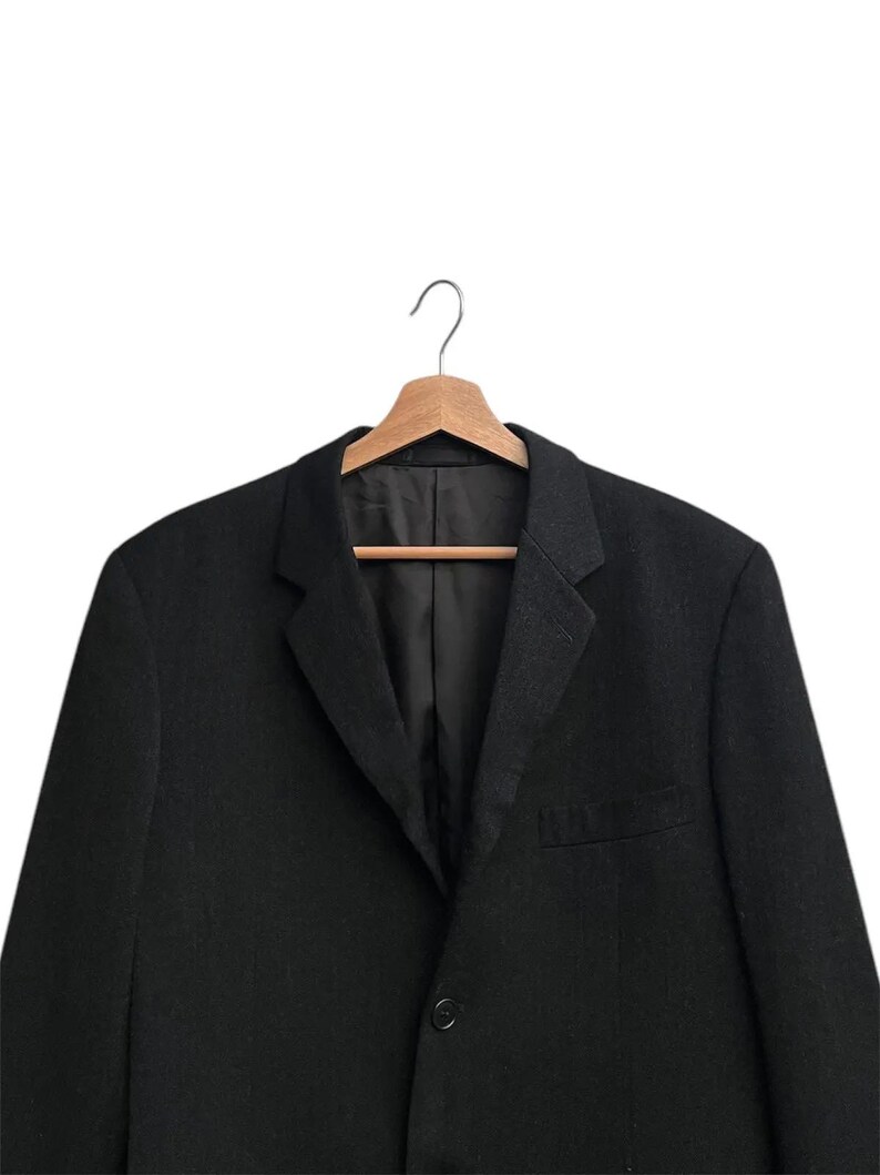 K&ouml;nnte beinhalten: Ein schwarzer Blazer h&auml;ngt an einem Holzkleiderb&uuml;gel. Der Blazer hat ein fallendes Revers, einen Ein-Knopf-Verschluss und eine Brusttasche. Der Stoff ist einfarbig dunkel und das Kleidungsst&uuml;ck ist ma&szlig;geschneidert.