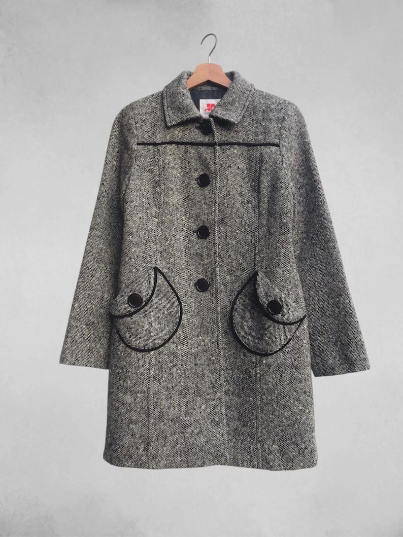 May include: A gray tweed coat with black trim and buttons. The coat has a classic design with a collar, long sleeves, and two decorative pockets. The coat is hanging on a wooden hanger.