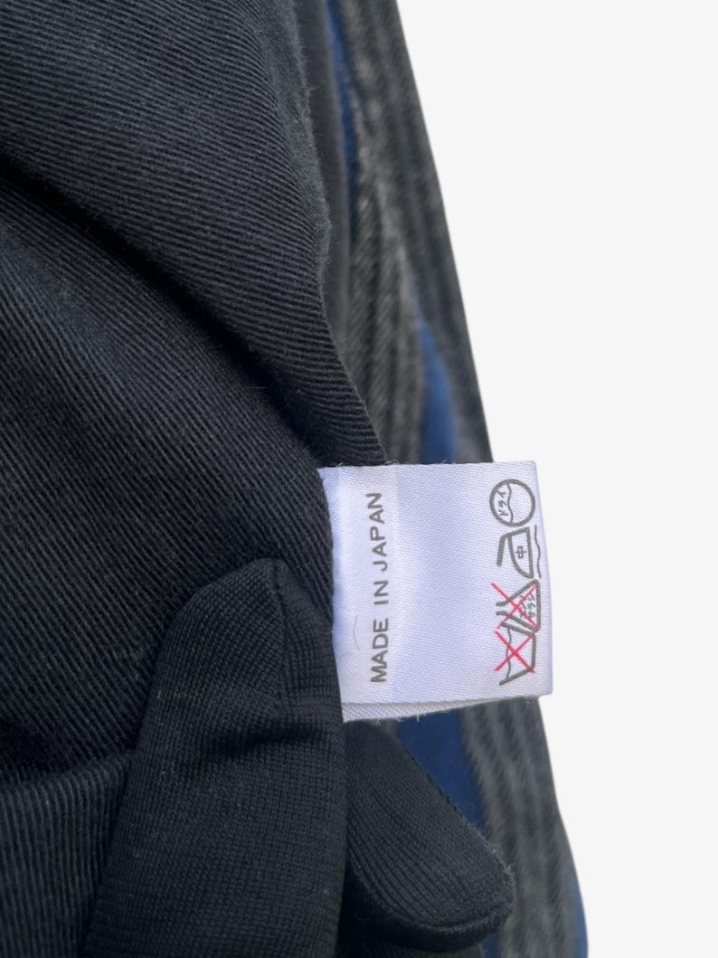 May include: Close-up of a black fabric with a blue stripe and a white tag. The tag reads "MADE IN JAPAN" in black text. Washing instructions are also printed on the tag.