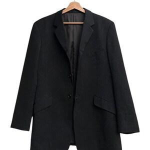 K&ouml;nnte beinhalten: Ein schwarzer Blazer h&auml;ngt an einem Holzkleiderb&uuml;gel. Der Blazer hat ein klassisches Design mit zwei Vordertaschen, einem Revers und einem Zwei-Knopf-Verschluss. Das Kleidungsst&uuml;ck ist aus einem einfarbigen, dunklen Stoff gefertigt.