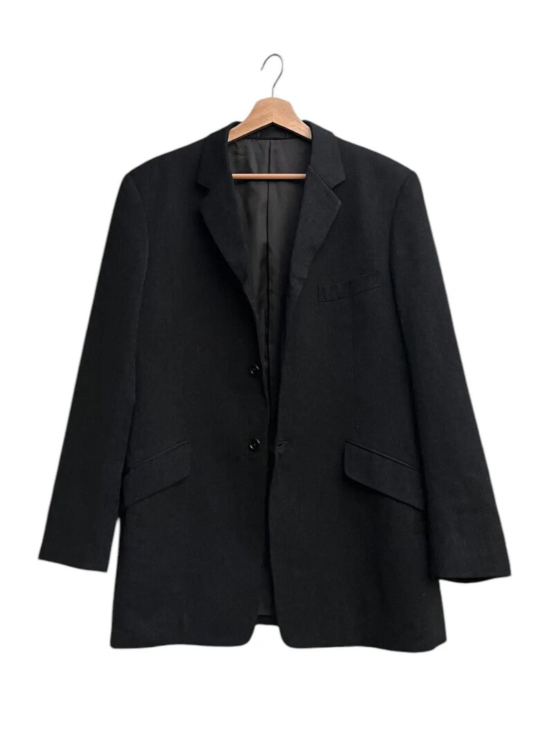 K&ouml;nnte beinhalten: Ein schwarzer Blazer h&auml;ngt an einem Holzkleiderb&uuml;gel. Der Blazer hat ein klassisches Design mit zwei Vordertaschen, einem Revers und einem Zwei-Knopf-Verschluss. Das Kleidungsst&uuml;ck ist aus einem einfarbigen, dunklen Stoff gefertigt.