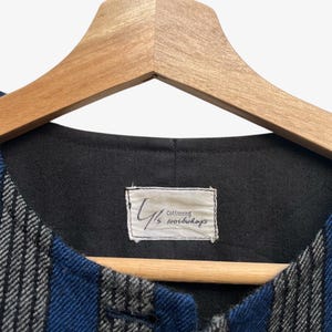 May include: A blue and gray striped vest with a black interior, hanging on a wooden hanger. The vest has a label that reads "Cottoning" and other text. The fabric has a textured appearance.