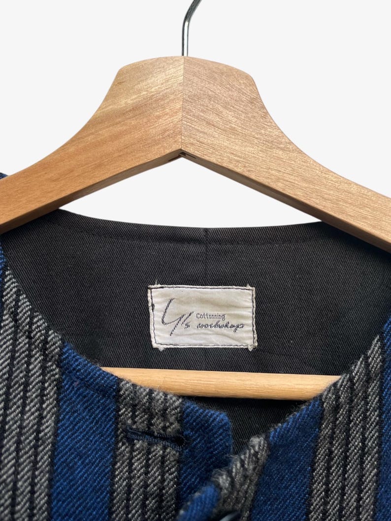 May include: A blue and gray striped vest with a black interior, hanging on a wooden hanger. The vest has a label that reads "Cottoning" and other text. The fabric has a textured appearance.