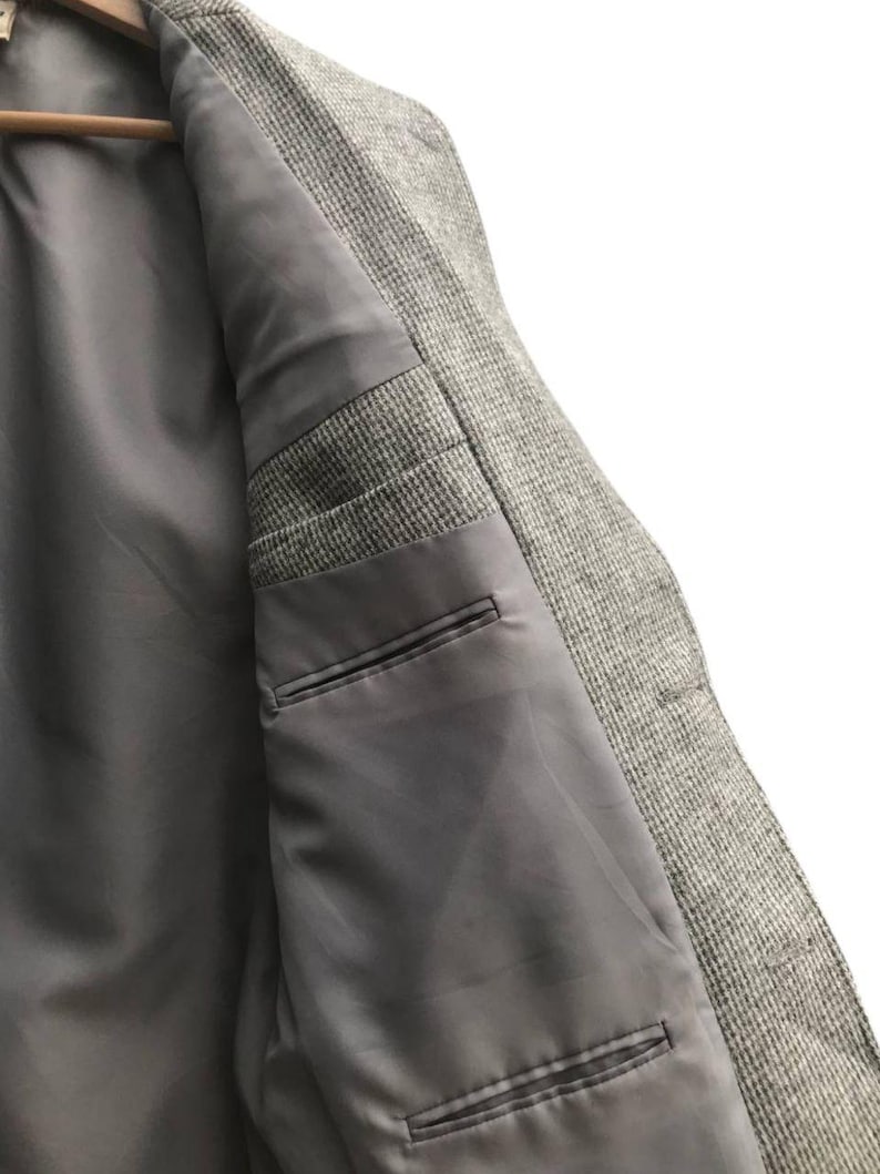 May include: Close-up of the inside of a gray tweed blazer. The interior features a smooth, gray lining with two pockets. The blazer is hanging on a wooden hanger, showcasing the tailored construction and quality of the garment.