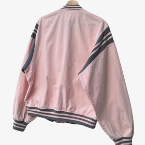 Puede incluir: Chaqueta bomber rosa con rayas grises en las mangas, el cuello y los pu&ntilde;os. La chaqueta est&aacute; colgada de una percha de madera sobre un fondo blanco. La chaqueta tiene un dise&ntilde;o cl&aacute;sico de bomber.