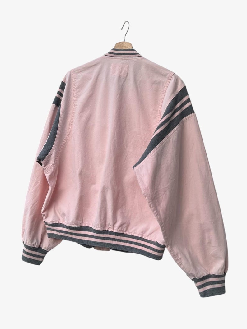 Puede incluir: Chaqueta bomber rosa con rayas grises en las mangas, el cuello y los pu&ntilde;os. La chaqueta est&aacute; colgada de una percha de madera sobre un fondo blanco. La chaqueta tiene un dise&ntilde;o cl&aacute;sico de bomber.