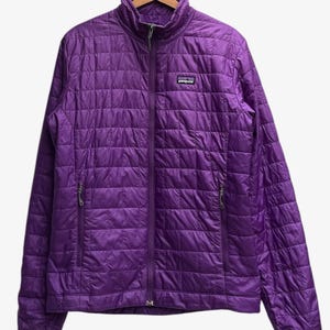 Puede incluir: Una chaqueta acolchada de color morado vibrante de la marca Patagonia con cremallera frontal completa, cuello alzado y dos bolsillos laterales con cremallera. La prenda presenta un patrón de costuras en cuadrícula y un logo en el pecho.