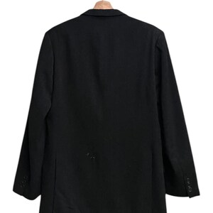 K&ouml;nnte beinhalten: Ein schwarzer Blazer h&auml;ngt an einem Holzb&uuml;gel. Der Blazer hat ein klassisches Design mit einem Zwei-Knopf-Verschluss und drei Kn&ouml;pfen an jedem &Auml;rmel. Der Stoff scheint eine einfarbige, dunkle Farbe zu haben.