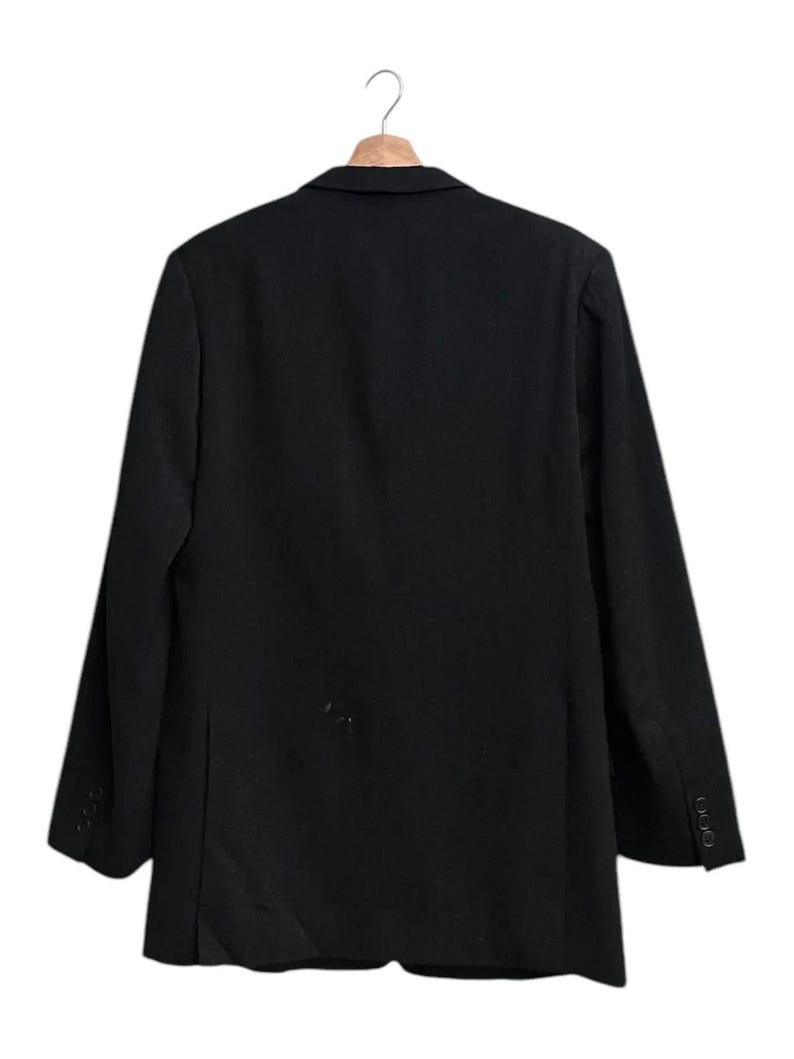 K&ouml;nnte beinhalten: Ein schwarzer Blazer h&auml;ngt an einem Holzb&uuml;gel. Der Blazer hat ein klassisches Design mit einem Zwei-Knopf-Verschluss und drei Kn&ouml;pfen an jedem &Auml;rmel. Der Stoff scheint eine einfarbige, dunkle Farbe zu haben.