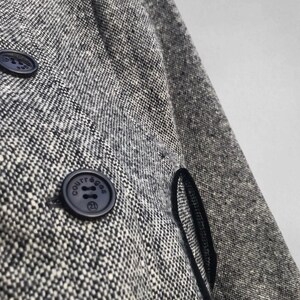 May include: Close-up of a gray and white tweed coat with two navy blue buttons. The buttons have the word "COURR&Egrave;GES" and the number "21" printed on them. The coat has a black trim along the edge of the pocket.