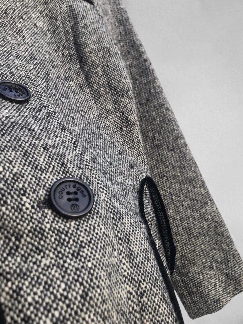 May include: Close-up of a gray and white tweed coat with two navy blue buttons. The buttons have the word "COURR&Egrave;GES" and the number "21" printed on them. The coat has a black trim along the edge of the pocket.