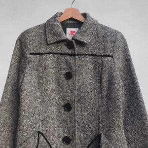 May include: A gray and white tweed coat with black trim and buttons. The coat has a classic design with a collar, three buttons down the front, and two decorative pockets. The coat is hanging on a wooden hanger.