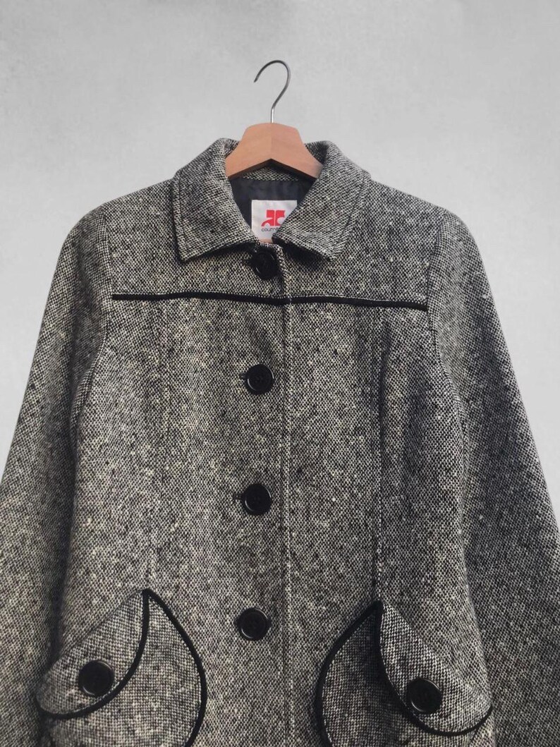 May include: A gray and white tweed coat with black trim and buttons. The coat has a classic design with a collar, three buttons down the front, and two decorative pockets. The coat is hanging on a wooden hanger.