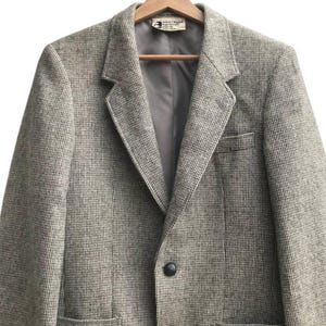 May include: A tweed blazer in shades of gray and brown hangs on a wooden hanger. The jacket features a notched lapel, two front pockets, and two black buttons. The interior lining is a solid gray color.