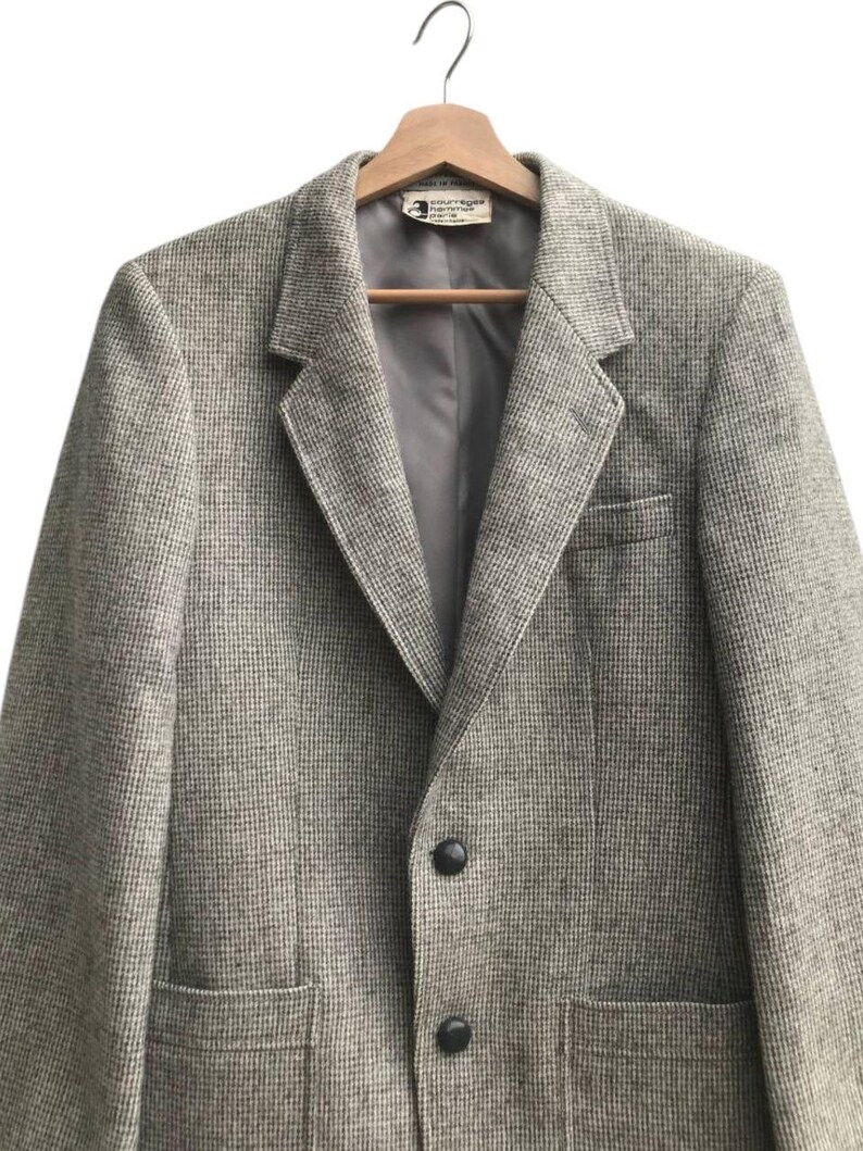 May include: A tweed blazer in shades of gray and brown hangs on a wooden hanger. The jacket features a notched lapel, two front pockets, and two black buttons. The interior lining is a solid gray color.