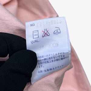 Puede incluir: Primer plano de una tela rosa con una etiqueta blanca. La etiqueta tiene texto y s&iacute;mbolos, incluyendo "NO. CE13-6012" y "QUAL. 100%". La tela es sostenida por una mano enguantada. Tambi&eacute;n se ven instrucciones de lavado.