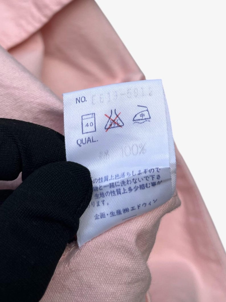 Puede incluir: Primer plano de una tela rosa con una etiqueta blanca. La etiqueta tiene texto y s&iacute;mbolos, incluyendo "NO. CE13-6012" y "QUAL. 100%". La tela es sostenida por una mano enguantada. Tambi&eacute;n se ven instrucciones de lavado.