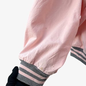 Puede incluir: Manga de chaqueta bomber rosa con pu&ntilde;o acanalado a rayas grises y rosas. La tela de la chaqueta es rosa claro y el pu&ntilde;o presenta tres rayas grises y rosas. La manga est&aacute; ligeramente arrugada.