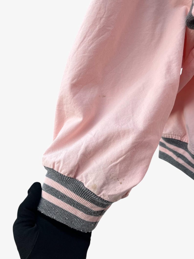 Puede incluir: Manga de chaqueta bomber rosa con pu&ntilde;o acanalado a rayas grises y rosas. La tela de la chaqueta es rosa claro y el pu&ntilde;o presenta tres rayas grises y rosas. La manga est&aacute; ligeramente arrugada.