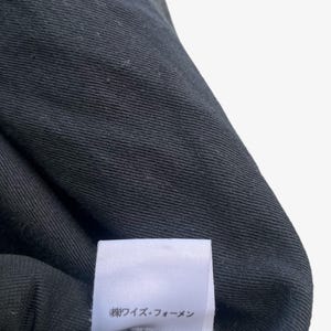 May include: Close-up of a black fabric with a textured weave. A white label with Japanese text is attached to the fabric. The fabric appears to be part of a garment, possibly a shirt or jacket.