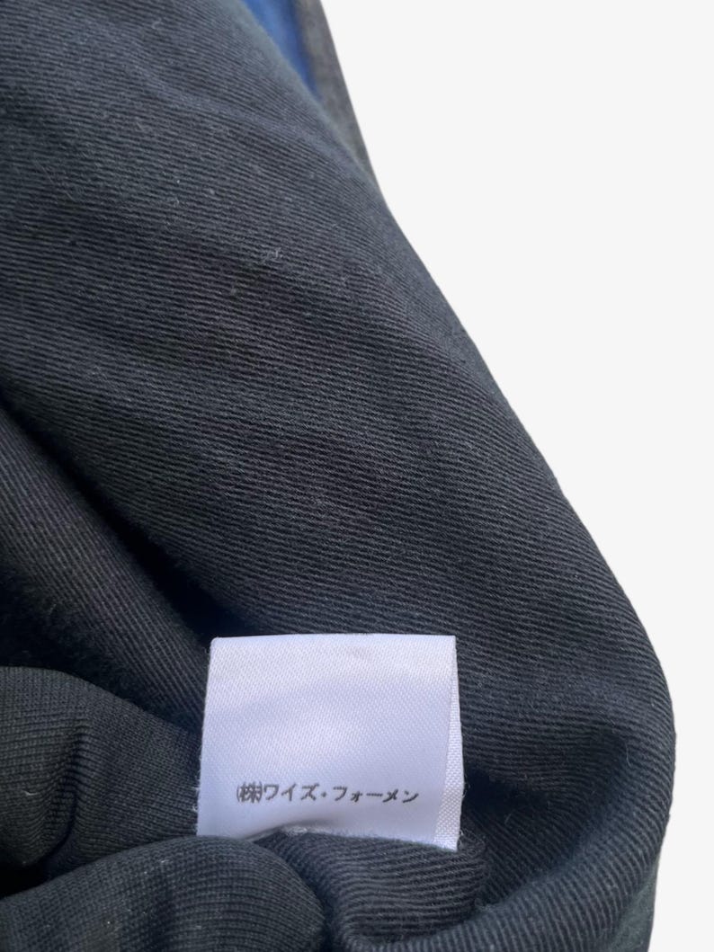 May include: Close-up of a black fabric with a textured weave. A white label with Japanese text is attached to the fabric. The fabric appears to be part of a garment, possibly a shirt or jacket.