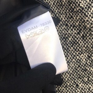May include: A white clothing tag with the text "EVDAM-78330" and care symbols. The tag is attached to a black garment lining, set against a black and white tweed fabric.