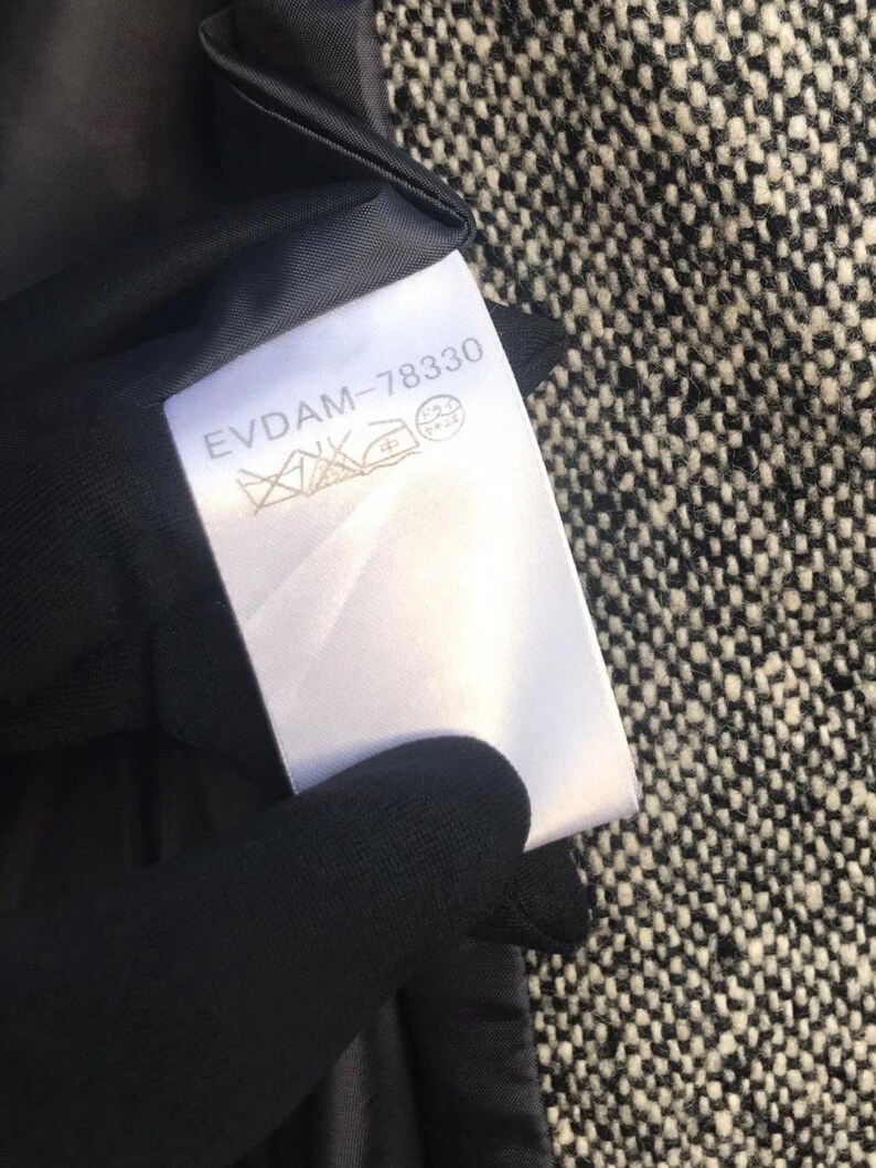May include: A white clothing tag with the text "EVDAM-78330" and care symbols. The tag is attached to a black garment lining, set against a black and white tweed fabric.
