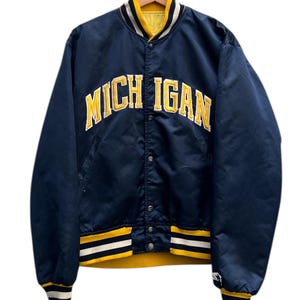 Peut inclure: Veste universitaire en satin bleu marine avec le mot "MICHIGAN" en lettres jaunes sur la poitrine. La veste a un col et des poignets jaunes avec des rayures blanches et jaunes. La veste est boutonnée et suspendue à un cintre.
