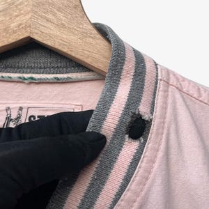 Puede incluir: Primer plano de un cuello de chaqueta rosa con ribete a rayas grises y rosas. La chaqueta est&aacute; abotonada con un bot&oacute;n negro. El cuello est&aacute; en una percha de madera.
